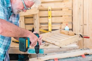 Senior man working with drill on a recycled pallet to make a chair. Hobby for retired people