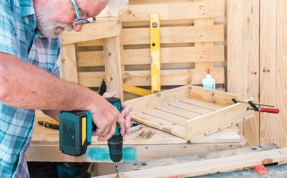 Senior man working with drill on a recycled pallet to make a chair. Hobby for retired people